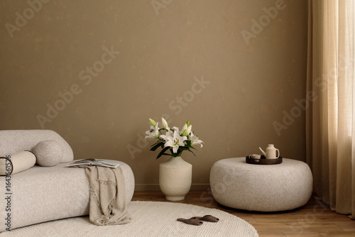 Fototapeta Naklejka Na Ścianę i Meble -  Elegant living space with beige sofa, large vase of white lilies, round pouf and textured neutral wall. Copy space.