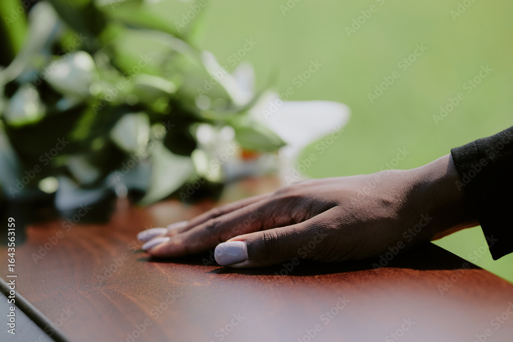 Obraz premium Black woman hand resting on wooden casket during funeral ceremony, blurred green background and floral arrangement visible, close up capturing gesture of farewell and mourning
