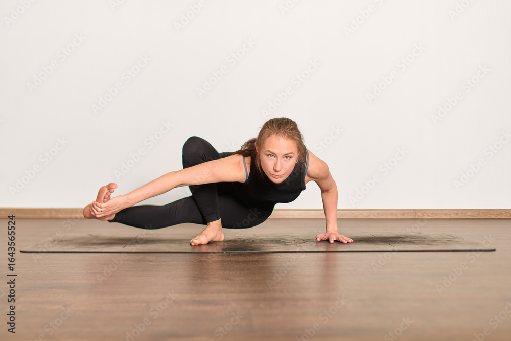 Obraz premium Woman performs advanced yoga balance pose on dark yoga mat. Minimalistic studio with wooden floor and white walls. Focused expression, natural light enhances serene atmosphere
