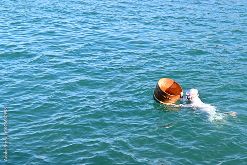 Ama diver in white attire swims with wooden tub in ocean.