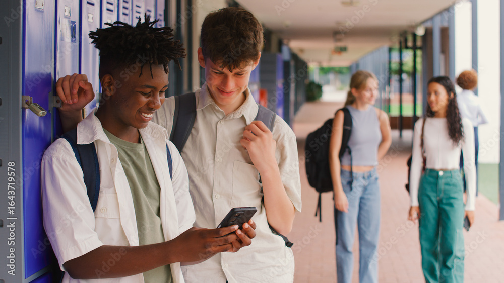 Obraz premium Two Male High School Or Secondary Students Looking At Social Media Or Internet On Phone By Lockers