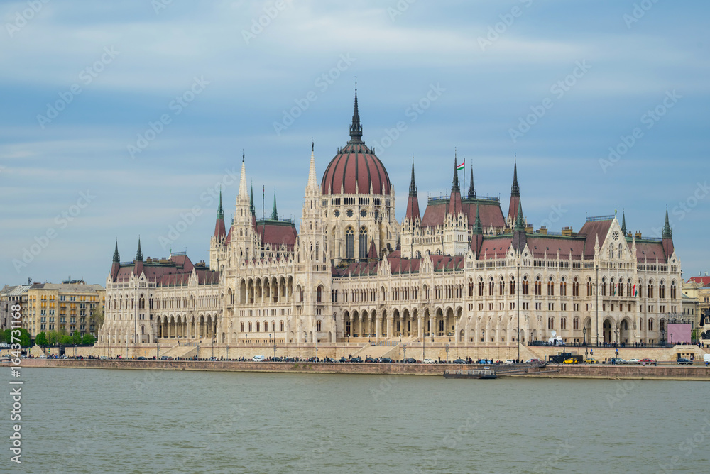 Fototapeta premium Stunning architecture of the Hungarian parliament building reflects in the Danube River. Visitors admire the landmark under a clear sky while boats glide past in the waterway.