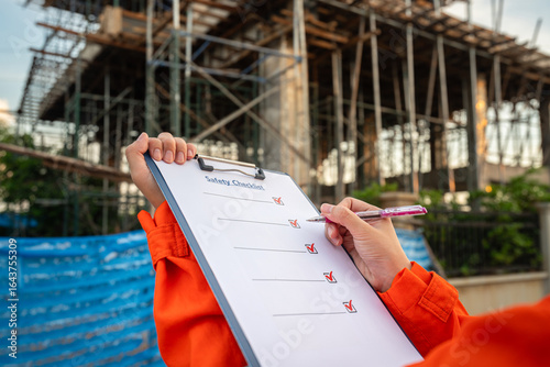 Action of an engineer is checkling on safety checklist form at construction working site (with background of house builing). Industrial safe work practice concept, close-up with selective focus.
