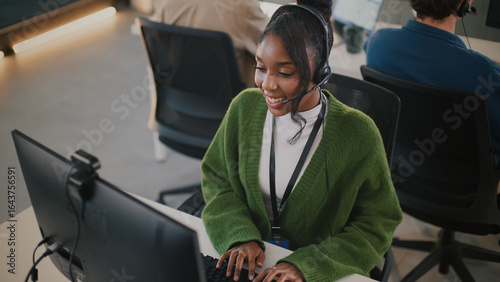 Smiling customer service representative wearing green sweater and headset works at computer in modern office environment, with colleagues in background