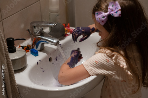 A little white girl washes her hands, which are heavily stained with paint, in the sink. A girl washes her hands under the tap after painting with paints