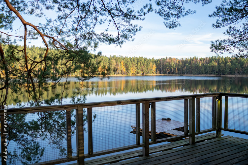 Naklejka premium Calm reflection of trees and sky on the lake at sunset from a wooden deck overlooking the water at a serene forest setting
