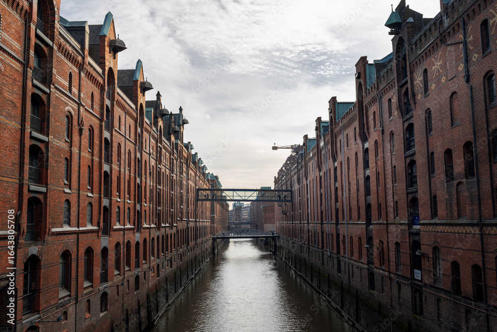 Naklejka premium Historical warehouse district in Hamburg features red brick buildings along a scenic canal under a cloudy sky