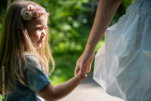 Фотография Outdoor half-length side view portrait of a blond little girl holding her mother’s hand, looking away thoughtfully, showing a tender and quiet moment