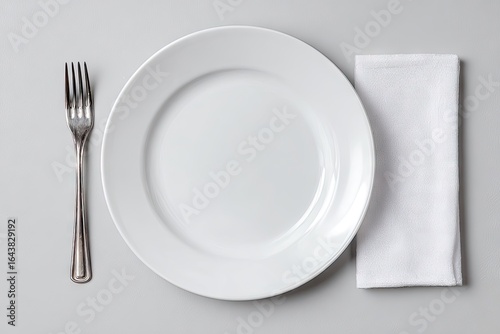Overhead view of a minimalist place setting a large white plate, a silver fork, and a white linen napkin on a light gray surface