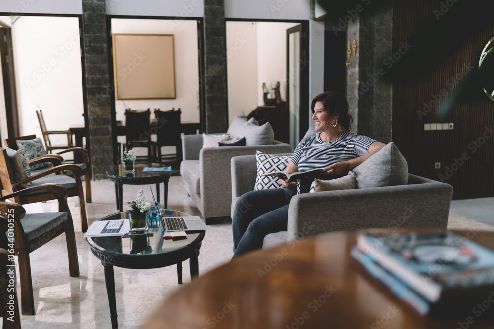 Fototapeta premium Freelancer smiling with book in hand, seated comfortably beside laptop, enjoying mindful moment of calm in digitally driven, coworking inspired interior.