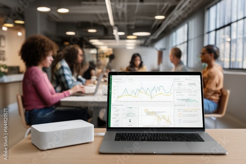 A laptop displaying data analytics sits on a desk in an open office. In the blurred background, four people work collaboratively, fostering a modern, dynamic environment