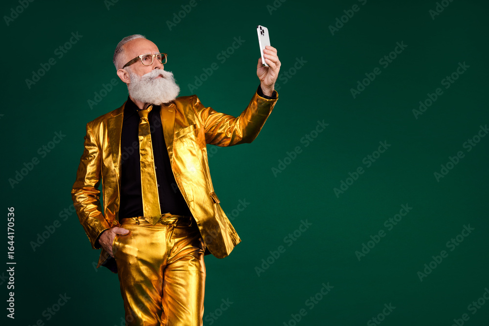 Fototapeta premium Elderly gentleman in glamorous golden suit taking a selfie on green background, embracing festive vibes of New Year's.