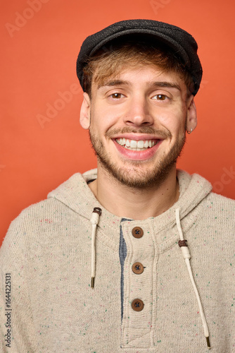 Close-up of smiling young Caucasian man in beige hoodie and flat cap, orange background