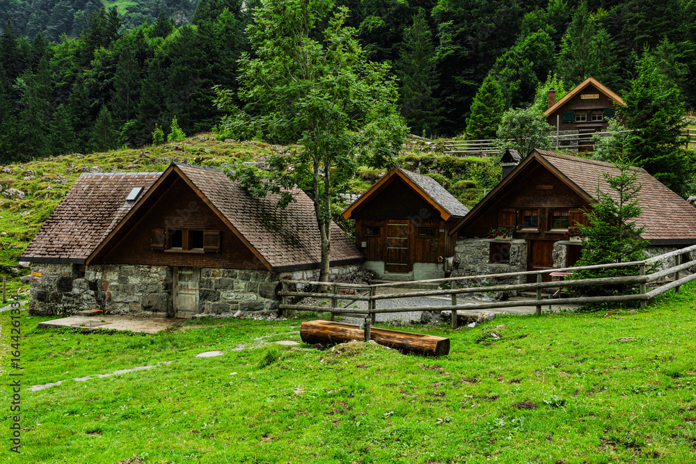 Obraz premium Traditional wooden and stone alpine houses set against a backdrop of lush forest and towering mountains near Seealpsee in the Appenzell Alps, Switzerland.