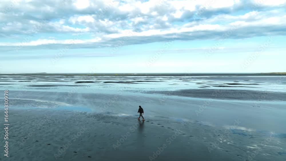 A person walks on a black sand beach near Vestrahorn mountain in Iceland. Sharp peaks rise in the background under a partly cloudy sky. A person walks on a black sand beach near Vestrahorn mountain in Iceland. Sharp peaks rise in the background under a partly cloudy sky.