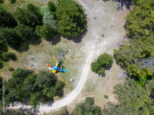Aerial view of a small playground area surrounded by trees and greenery, featuring slides and climbing structures, located along a dirt path in a natural forest setting, perfect for family outings and