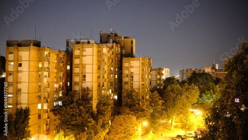 Night view of residential buildings with warm glowing windows, surrounded by trees and streetlights. Urban cityscape and life at night.