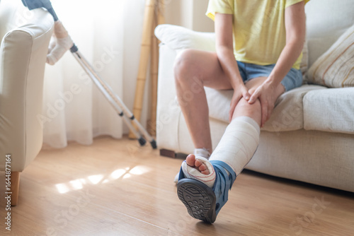 Patient wearing an orthopedic boot, with crutches nearby, massaging their injured leg while sitting on a sofa, recovering from a metatarsal fracture