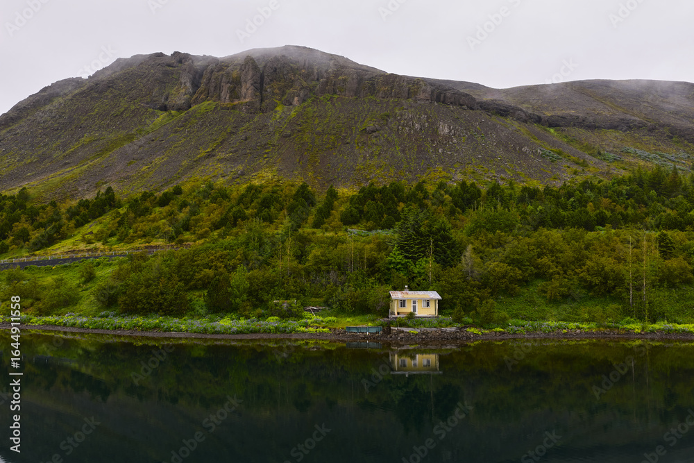 Fototapeta premium Small yellow house reflecting in a lake with green trees and mountain