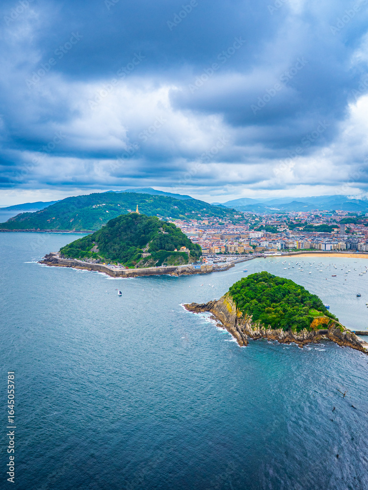 Naklejka premium Scenic view of La Concha Bay in San Sebastian, Spain, featuring Santa Clara Island, Mount Urgull, dramatic clouds, lush hills, city skyline, and sandy beach.