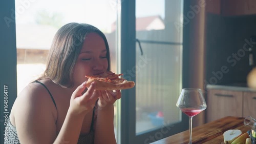 Elegant woman enjoying a slice of pizza with a glass of wine on a rustic table, captured in warm light, sharing good food with a friend in a cozy kitchen setting