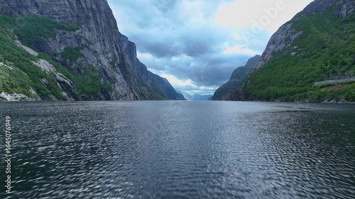 A drone glides over a fjord in Lysebotn, showcasing steep cliffs with greenery, calm reflective water, and dramatic peaks under a cloudy sky.