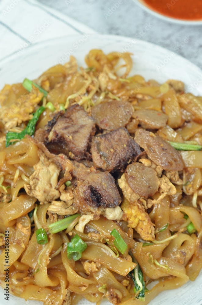 Char Kway Teow, fried rice flat noodles with beef, egg, and vegetables on a white plate, Indonesian dish. Char Kway Teow, fried rice flat noodles with beef, egg, and vegetables on a white plate, Indonesian dish.