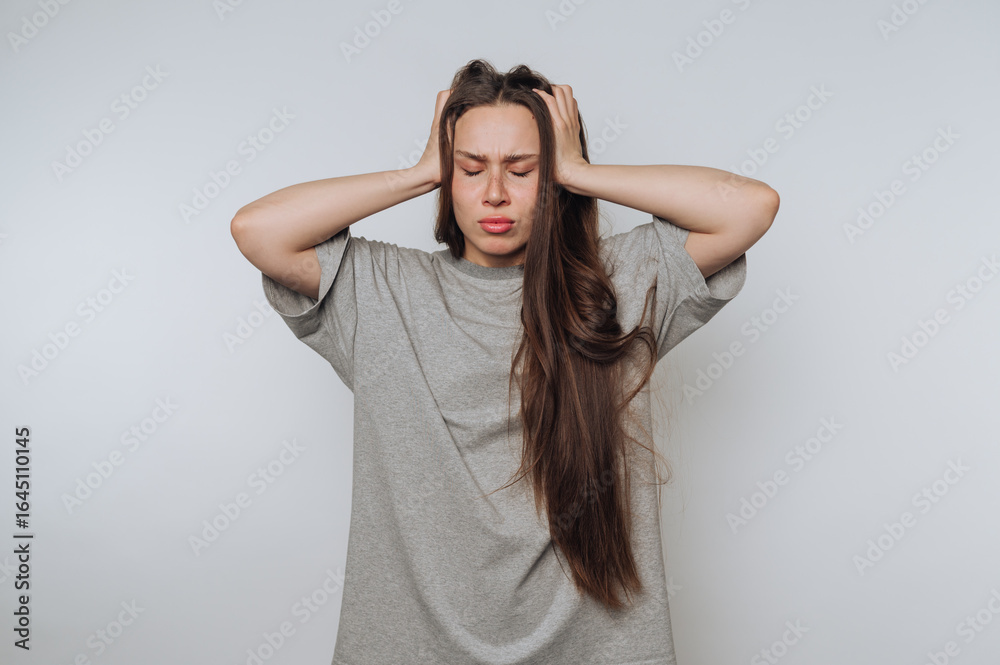 Fototapeta premium portrait of young frustrated woman holds her head in both hands, depicting clear signs of stress on light background