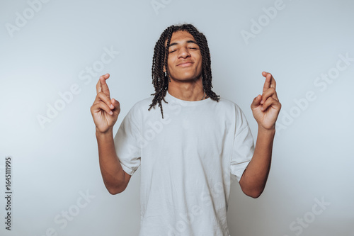 portrait of young man smiles while crossing his fingers, symbolizing hope and wishing for good luck on light background