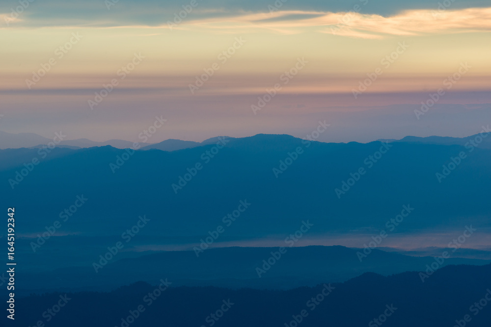 Obraz premium Sunset over misty layered mountains, evoking hope, reflection, and timeless beauty. Golden sunset casting warm light over layers of mountain ridges, with soft mist and pastel-colored sky 