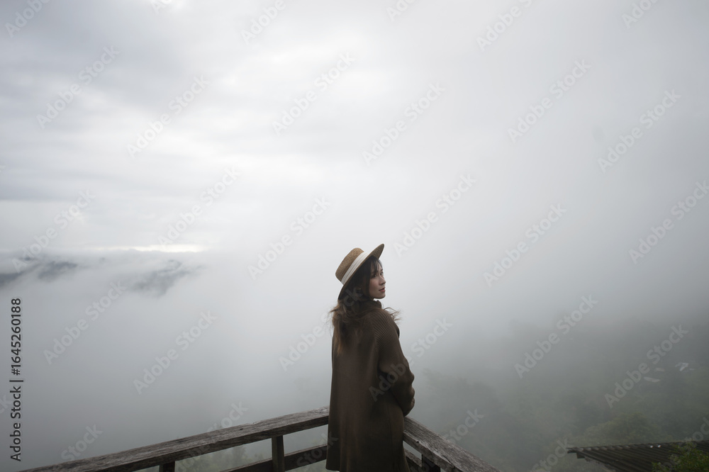 Fototapeta premium A woman stands on a wooden deck, gazing into a foggy landscape, enveloped by clouds and nature