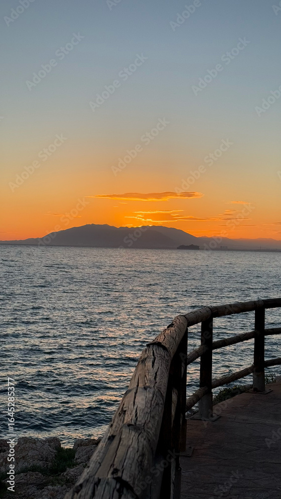 Malaga, Spain. Sunset on the Rincon de la Victoria, Malaga with views of the mountain El Monte Calamorro. Scenic sunset view over ocean with wooden railing and distant mountains in the background Malaga, Spain. Sunset on the Rincon de la Victoria, Malaga with views of the mountain El Monte Calamorro. Scenic sunset view over ocean with wooden railing and distant mountains in the background