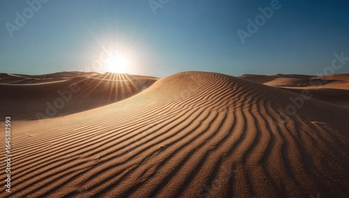 Sun-drenched sand dunes ripple gently across the desert landscape under a brilliant, clear sky; the sun's rays create a starburst effect