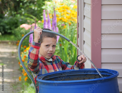 Young boy in a plaid shirt helps in the garden, filling a large blue rain barrel with water from a hose on a sunny August day