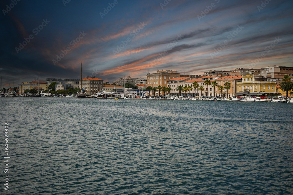 Obraz premium Colorful buildings lining zadar waterfront at sunset, with boats moored in the harbor and dramatic sky reflecting in the adriatic sea