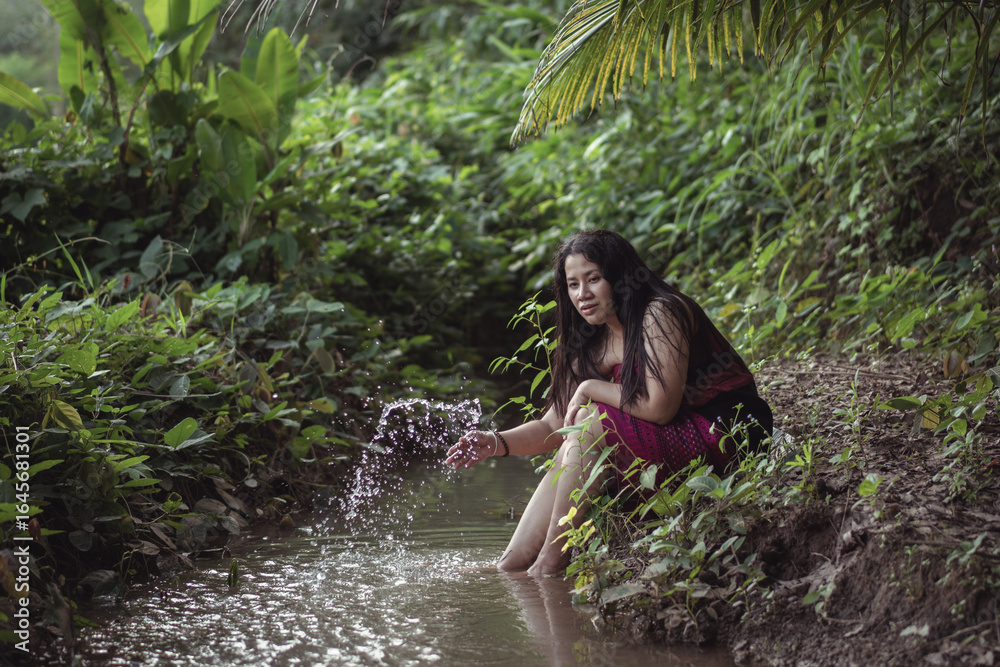 Fototapeta premium Serene Young Woman Sitting by a Stream in a Lush Green Forest, Enjoying Nature, Capturing the Essence of Tranquility and Peaceful Reflection