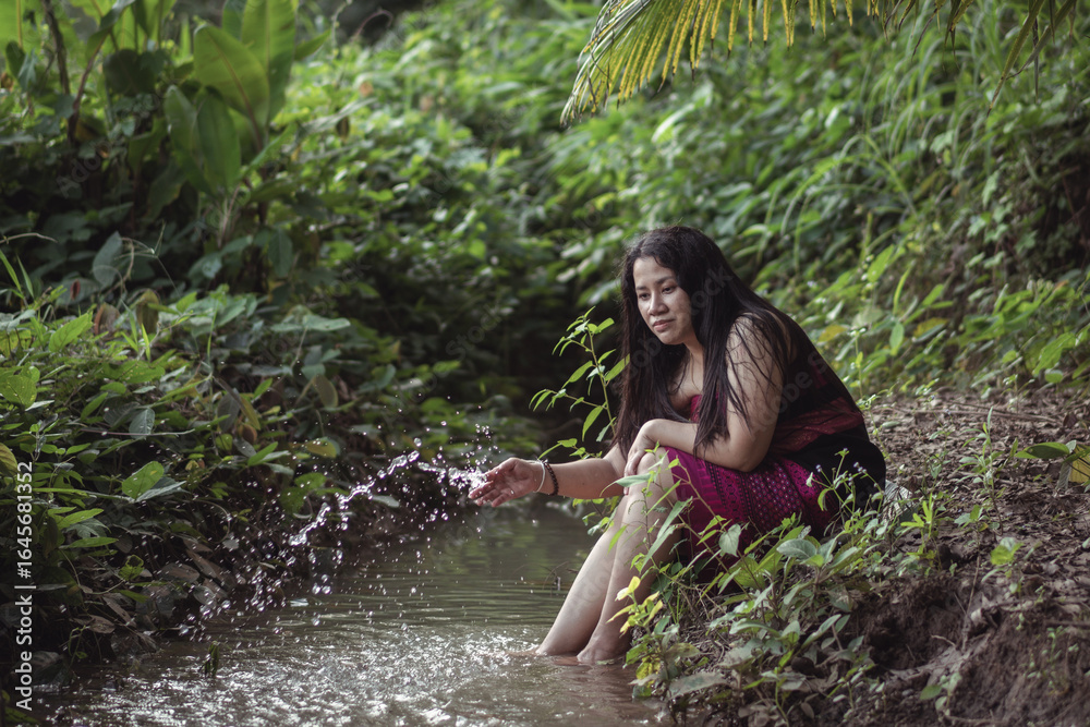 Fototapeta premium Young woman sitting by a serene stream in lush green foliage, gently splashing water while enjoying peaceful nature in a tropical setting