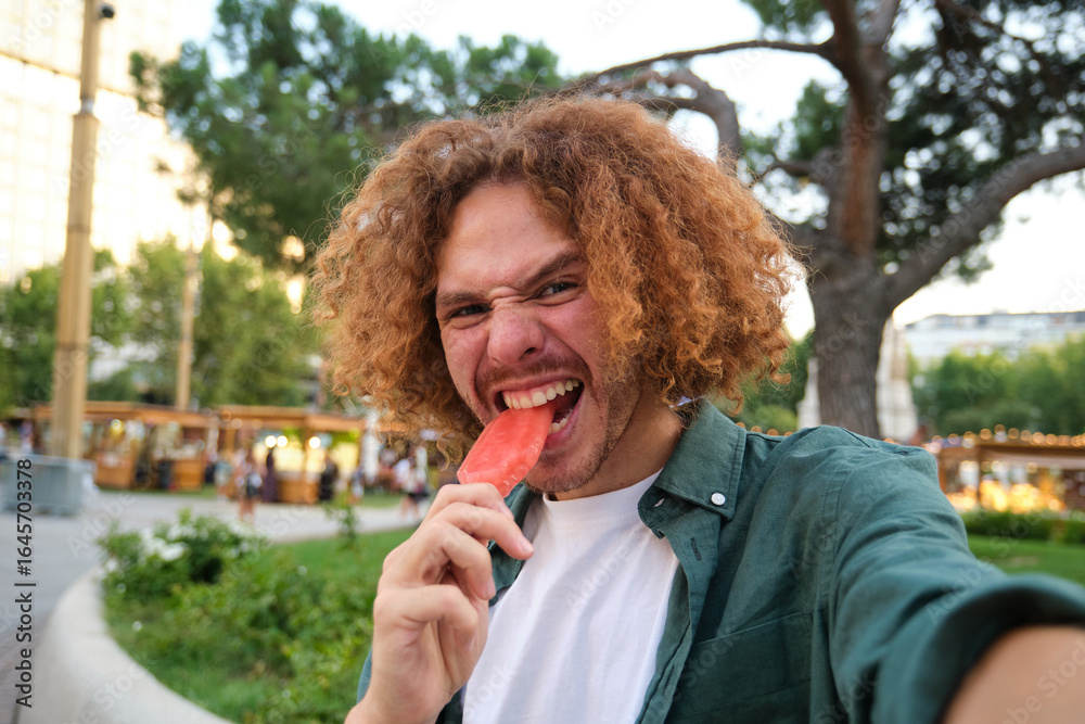 Fototapeta premium Ginger man taking a selfie while biting a popsicle in a park during summer
