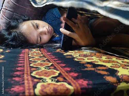 Canvastavla Little Asian girl lying on the bed while playing with her cell phone, covered in