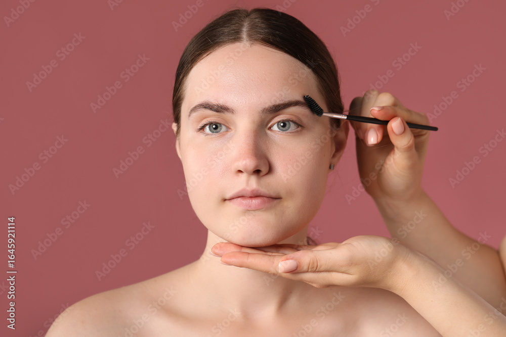 Fototapeta premium Cosmetologist brushing young woman's eyebrow with spoolie on pink background, closeup