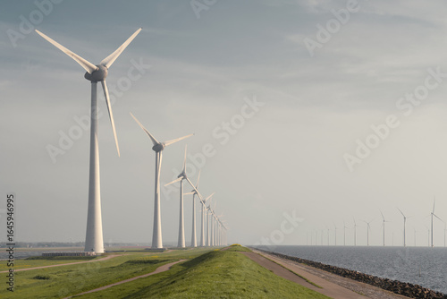 Wind turbines on green grass in Flevoland, Netherlands, with additional turbines faintly visible far offshore in the IJsselmeer. Captured on a sunny spring day with a clear blue sky.