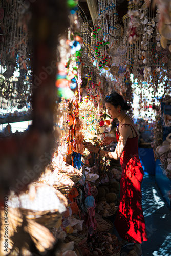 Woman in red dress shopping at a sunlit seaside shell market stall, surrounded by hanging seashell mobiles and coastal crafts; selective focus, natural light