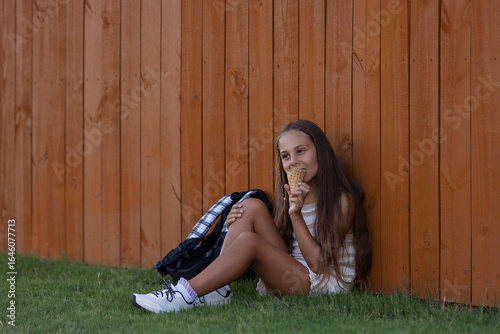 girl sitting on the grass by a wooden fence, eating an ice cream cone with a backback next to her.