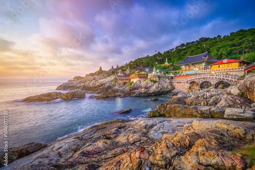 A scenic view of Haedong Yonggungsa Temple built on the rocky coastline in Busan, South Korea, during a colorful sunrise with ocean waves and dramatic sky.