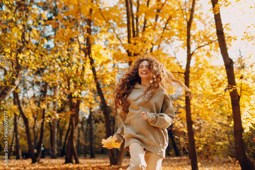 Smiling young woman plays and jumping dancing in the autumn park with the yellow leaves at sunset