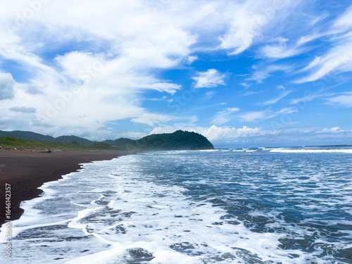 Wide view of Camaronal Beach in Costa Rica, with waves breaking on dark sand under a blue sky filled with clouds.