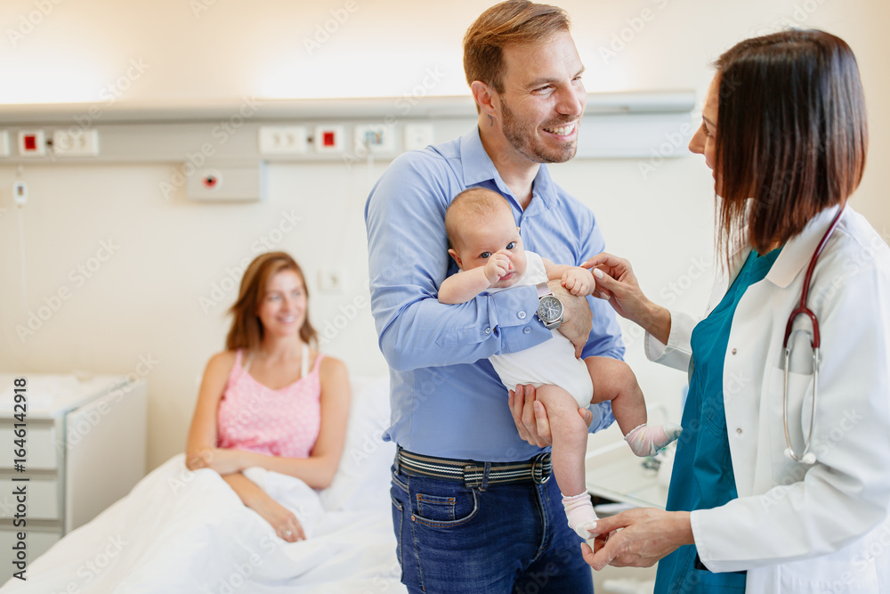 Fototapeta premium Smiling Father Holding Newborn with Doctor in Bright Hospital Room with Mother Resting Nearby