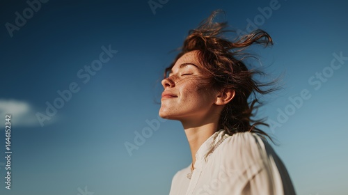 A woman with closed eyes enjoys the fresh air, hair blowing in the breeze under a clear blue sky.