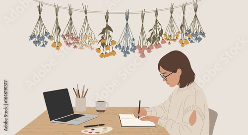 Woman writing in her journal at a wooden desk with laptop and flower decoration above creating an artistic and cozy atmosphere