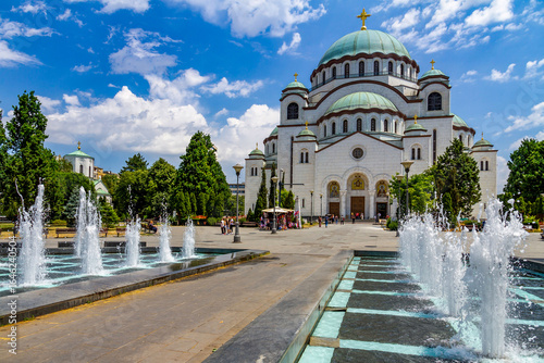 The Church of Saint Sava in Belgrade, a stunning example of Byzantine architecture, shines under a blue sky with white clouds. Greenery and fountains surround the square. Serbia
